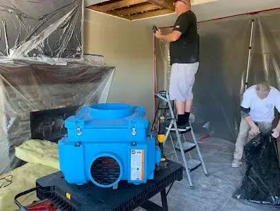 Crew opening a wall behind plastic containment with an air scrubber running during structural drying