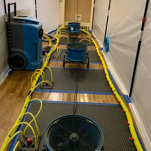 Hardwood floor drying mats with yellow hoses and a dehumidifier inside plastic containment